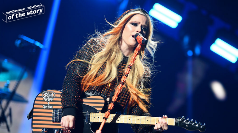 An image of Avril Lavigne holding a guitar in front of a microphone.